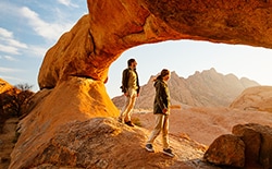 man and woman stand on rock formation and look out at mountainous horizon