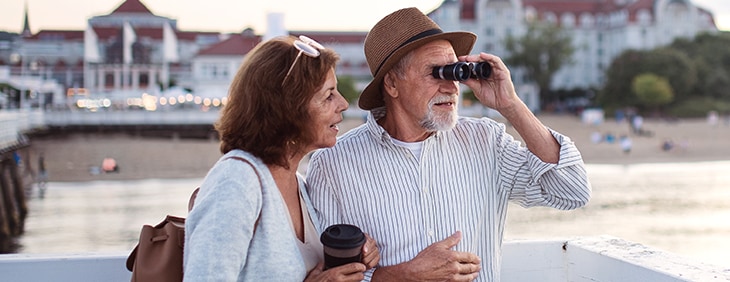 older couple stands on peer as man looks through binoculars 