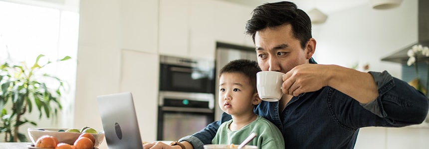 man drinks coffee and works at laptop while holding young son on lap