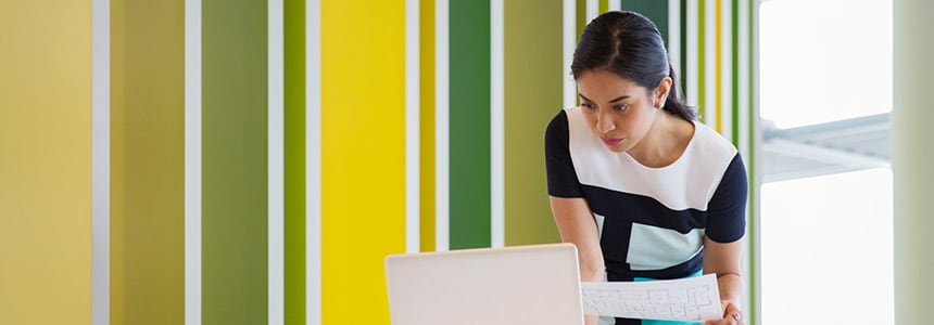 woman stands and works at laptop while holding document