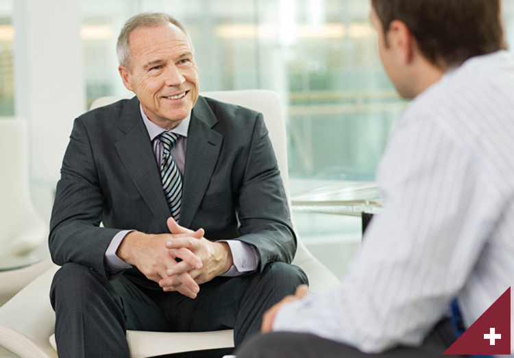 Older businessman sits in chair with hands clasped and smiles at younger businessman