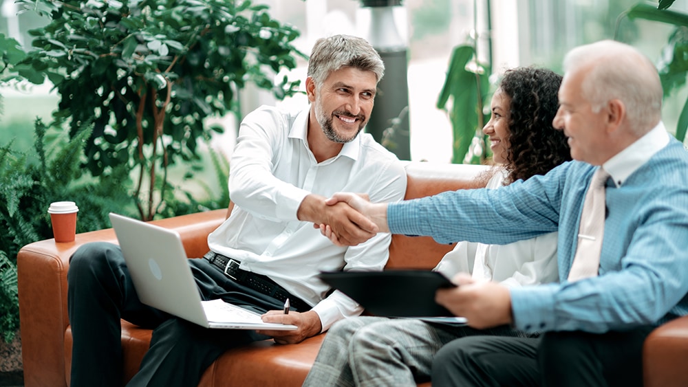 three people sit on couch with laptop and electronic tablet. The two farthest apart are shaking hands