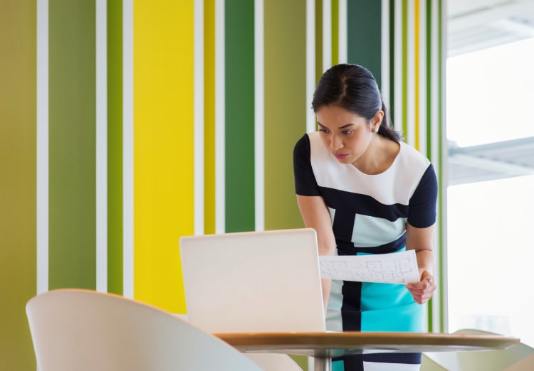 woman stands and works at laptop while holding document