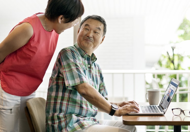 Man sits at desk with laptop while smiling at wife who leans over his shoulder