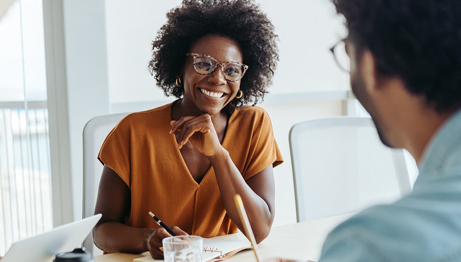 smiling woman sits at desk and and takes notes while talking to man