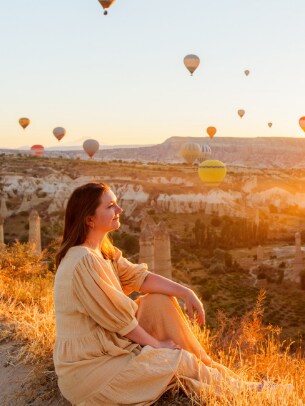 woman sits in field as hot air balloons raise into the sky in the distance