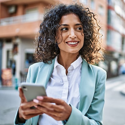 woman holds cell phone in front of her on city sidewalk and looks off to the right