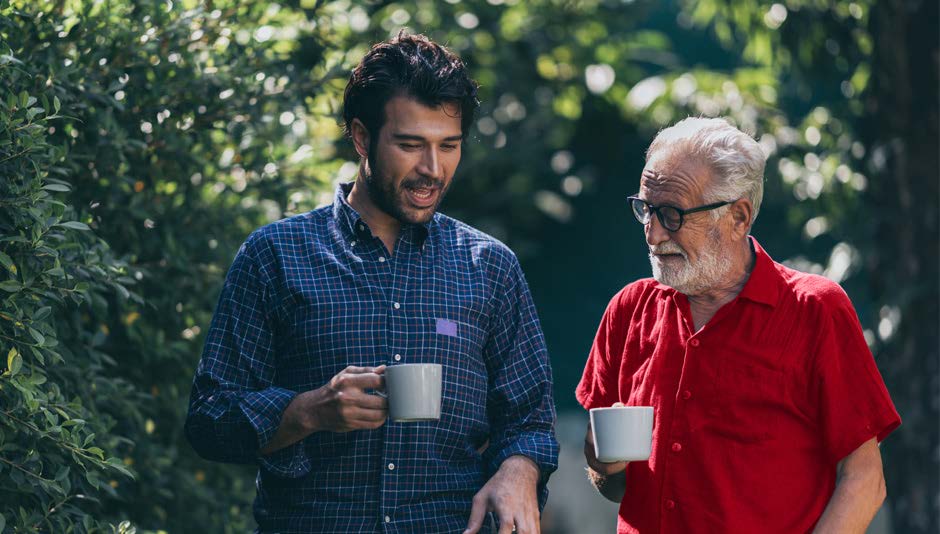 Two men walk in garden drinking from mugs