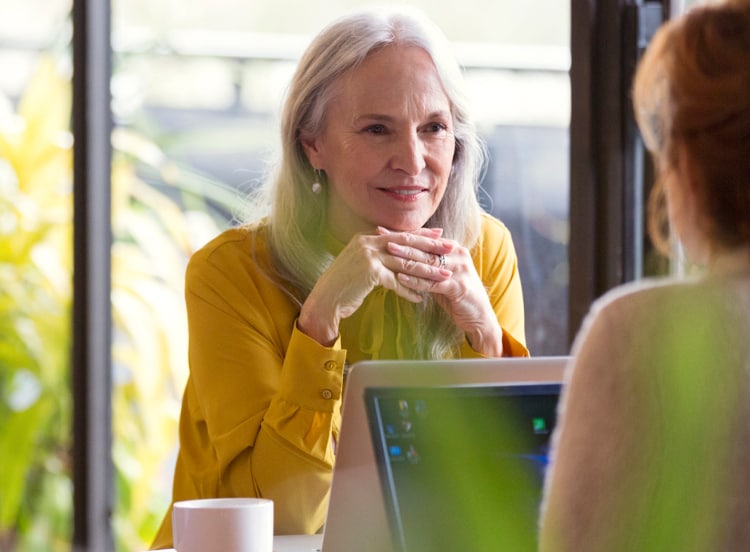 older woman sits at desk and looks across at another person