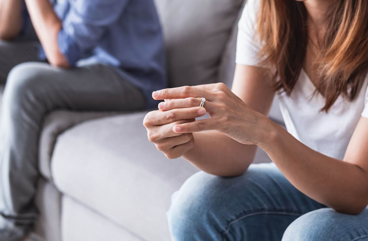 A maried couple are sitting at opposite ends of a couch. The woman is touching her wedding ring.