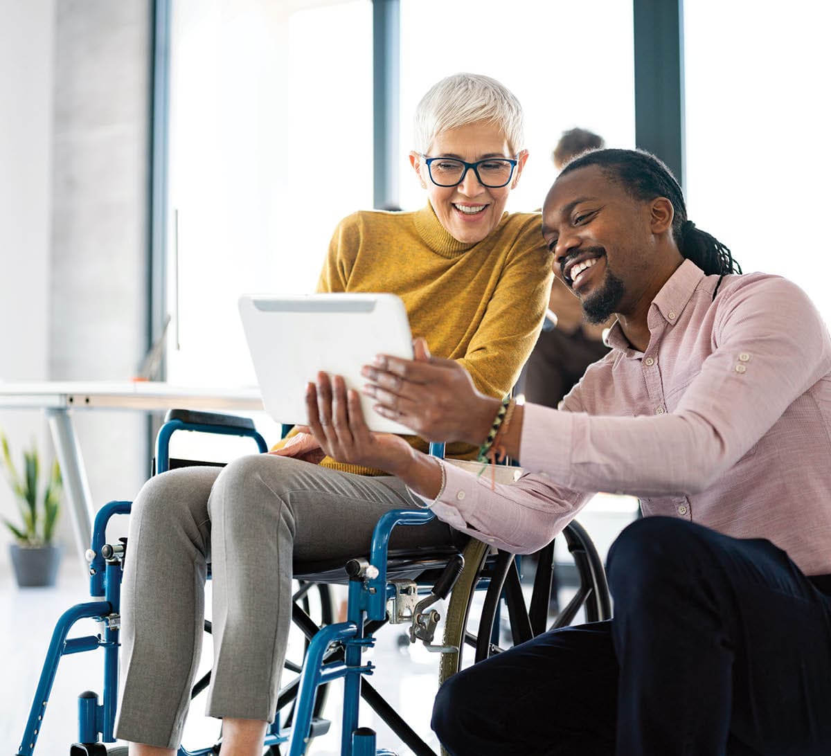 A man is kneeling next to a woman in a wheelchair. They are looking at an electronic tablet.