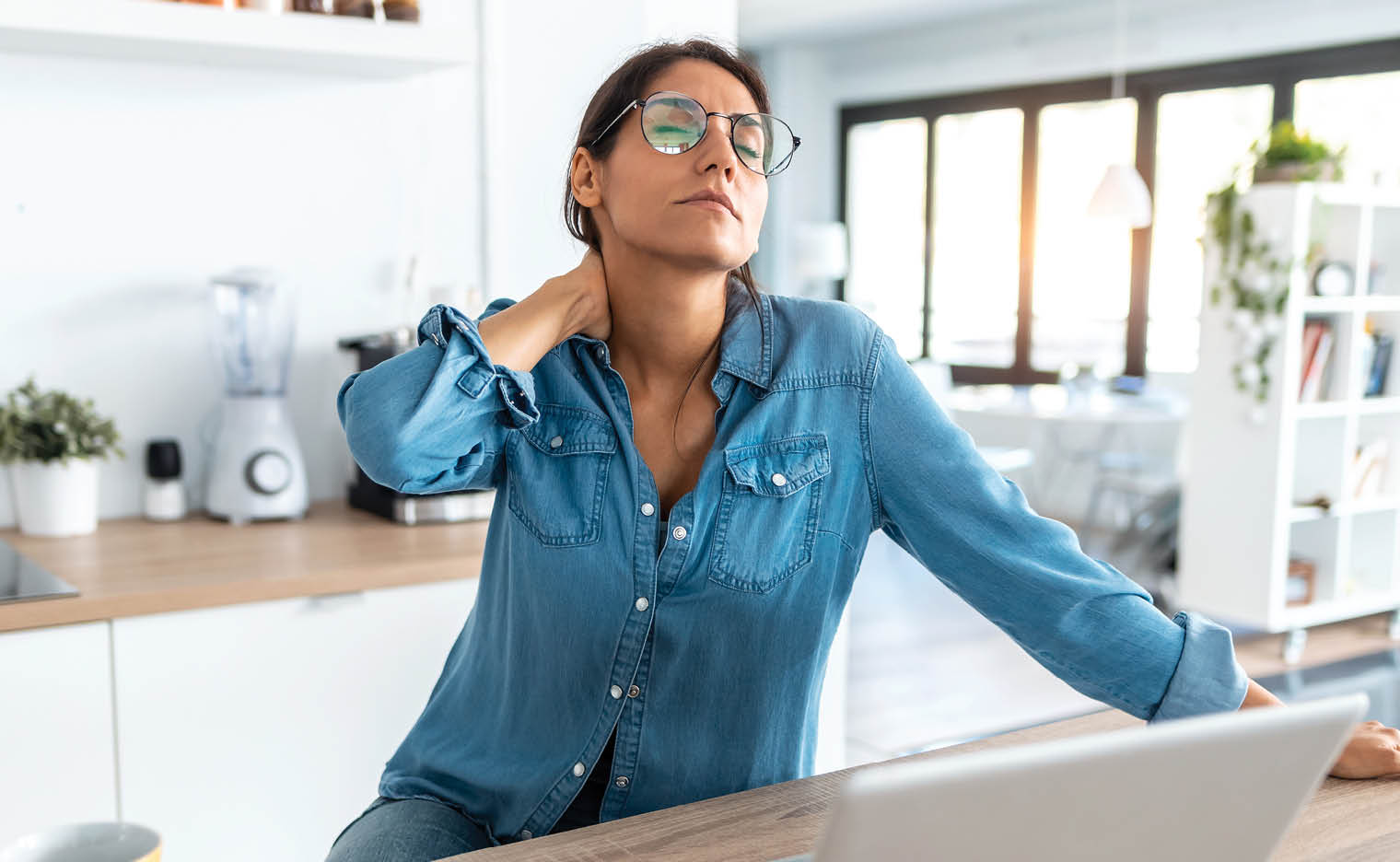 woman working on laptop looking stressed. she is holding the back of her neck and stretching.