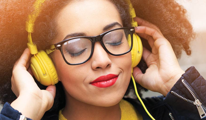 Attractive young woman listening music. Yellow headphones on head, eyeglasses, wear black jacket, yellow blouse. Hands on headphones, eyes closed.