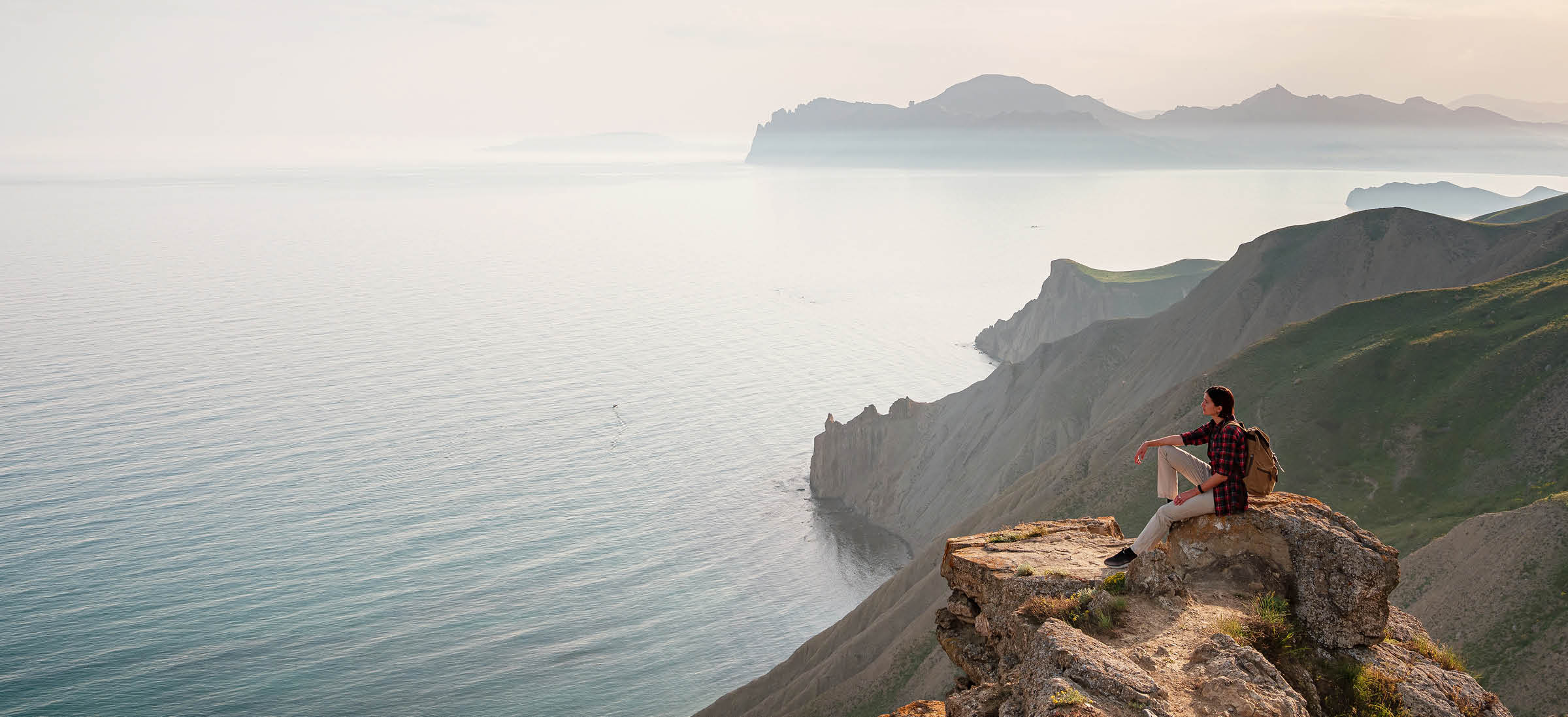 a hiker sits on mountaintop rocks looking out over vast water and mountains