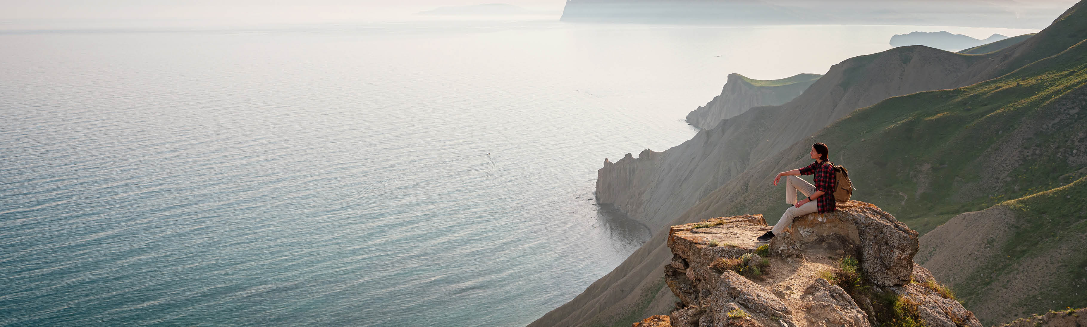 a hiker sits on mountaintop rocks looking out over vast water and mountains