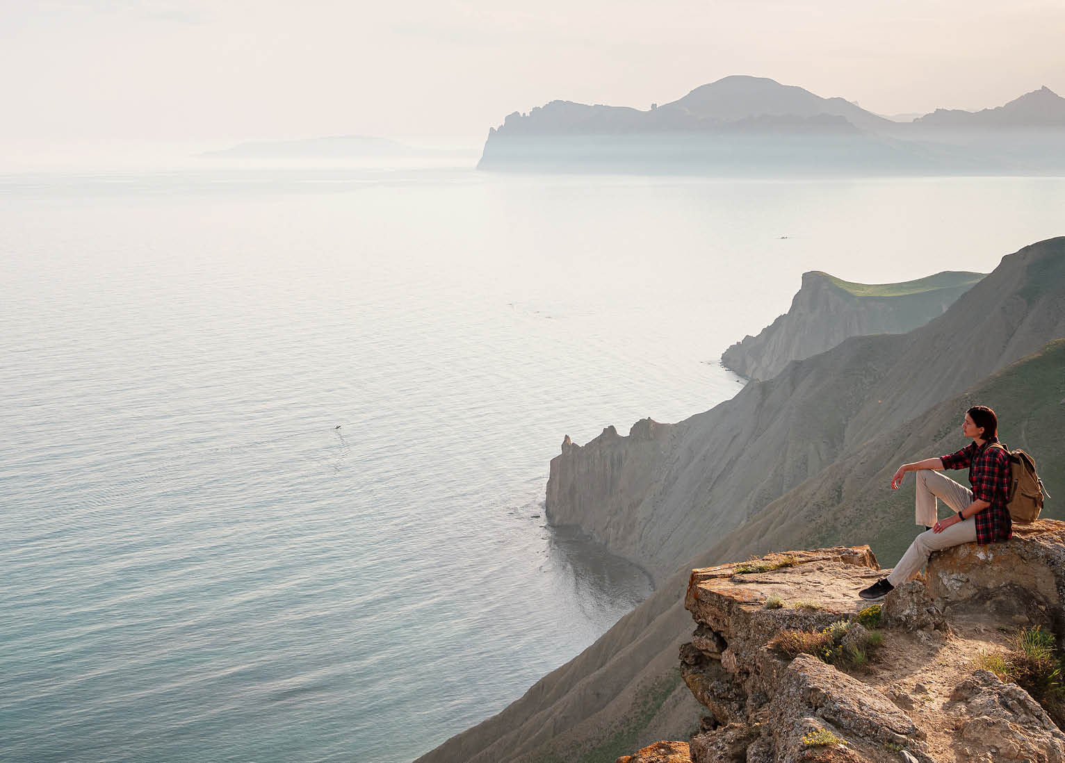 a hiker sits on mountaintop rocks looking out over vast water and mountains
