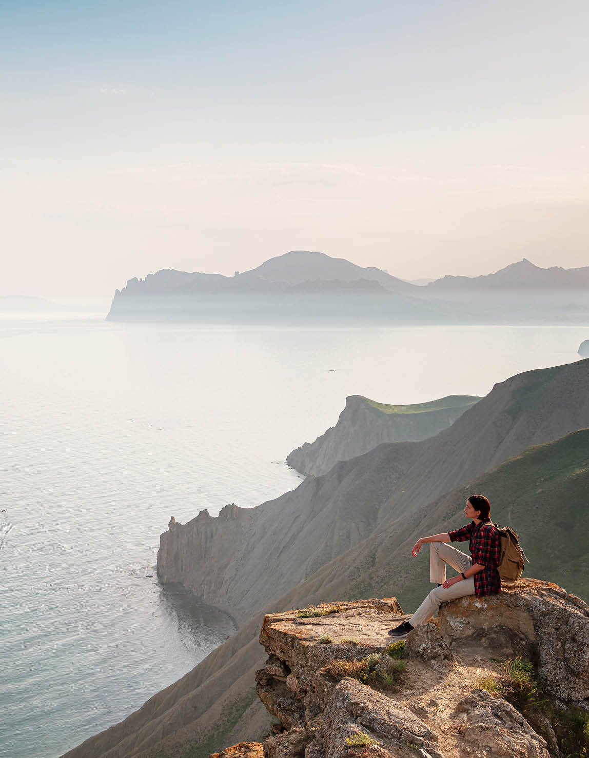 a hiker sits on mountaintop rocks looking out over vast water and mountains