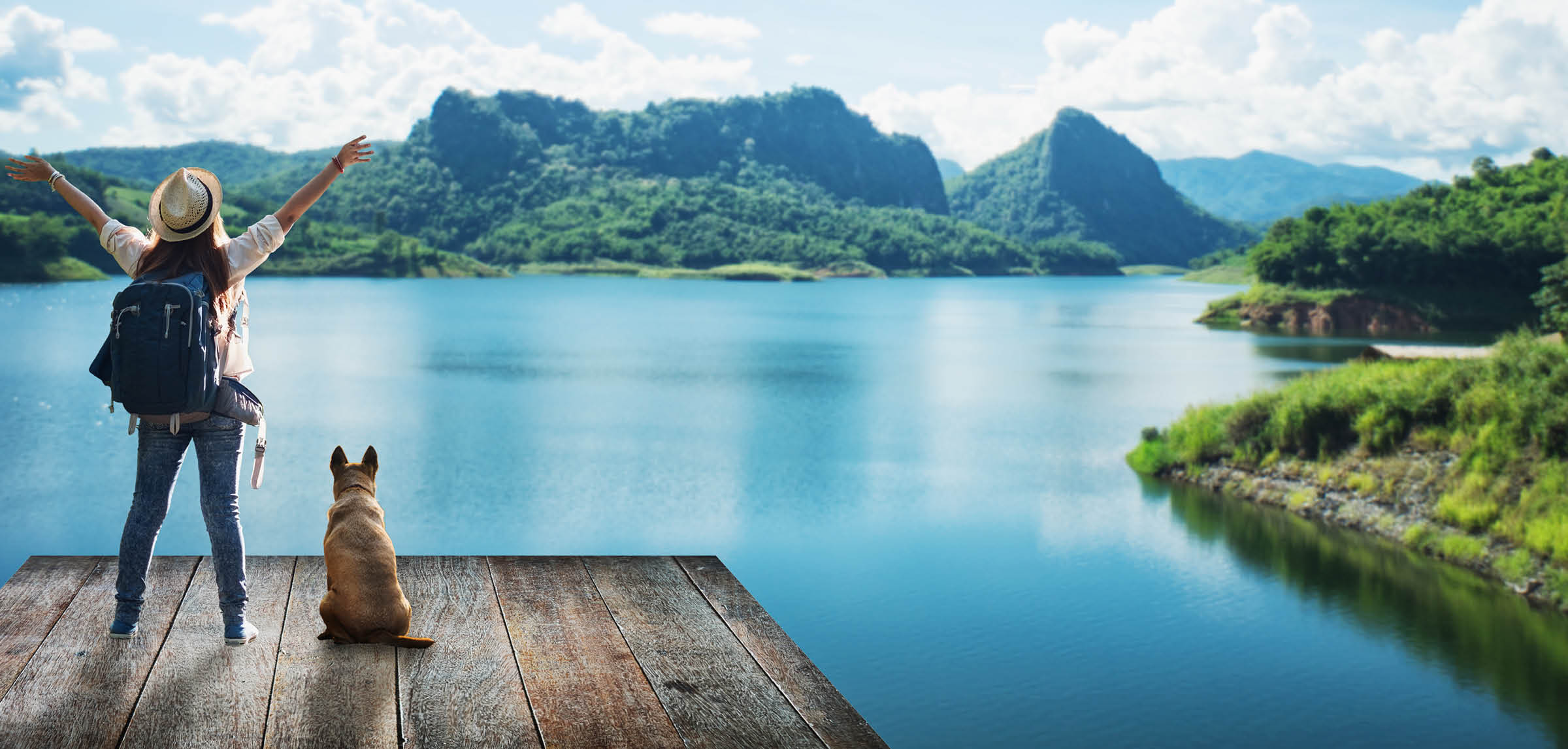 a woman stands with arm raised looking at mountains across water. a dog sits beside her.