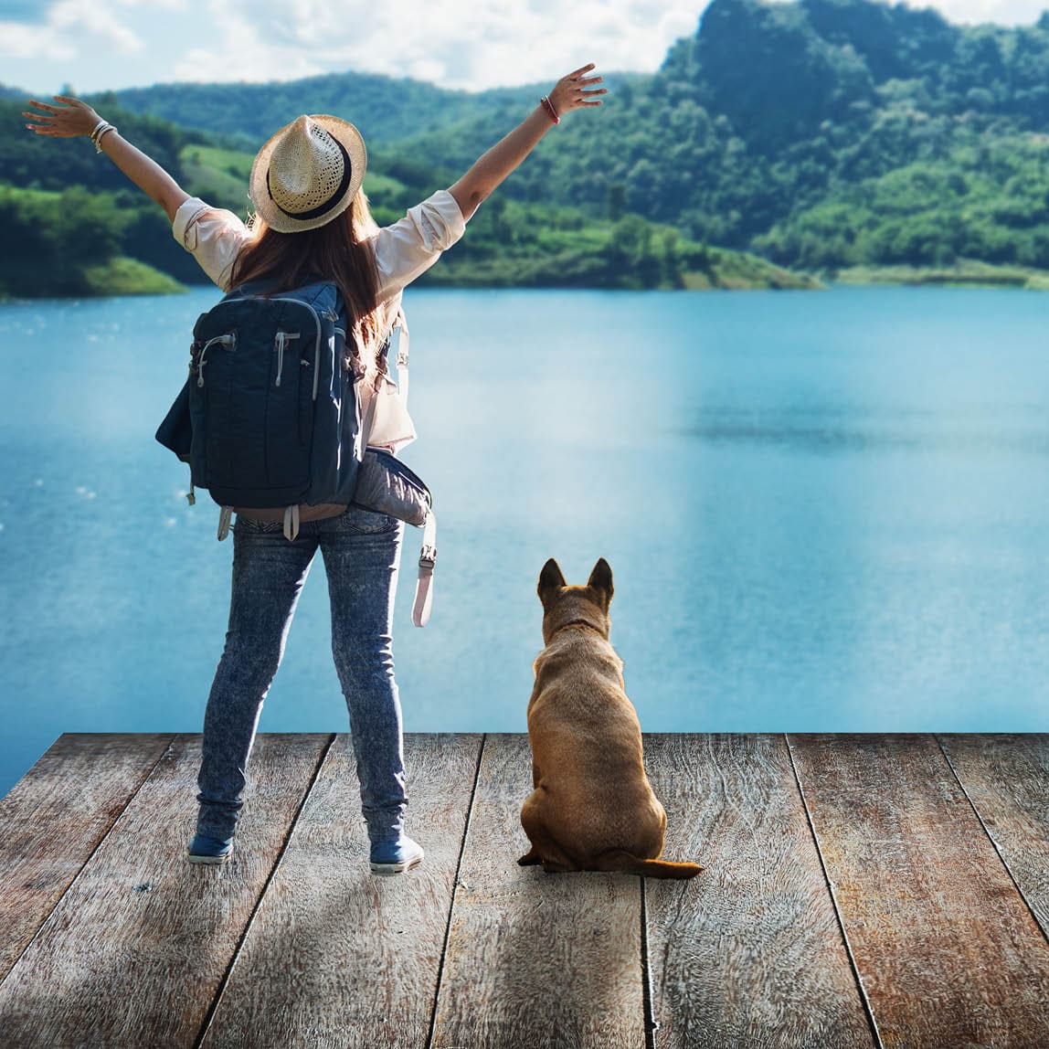 a woman stands with arm raised looking at mountains across water. a dog sits beside her.
