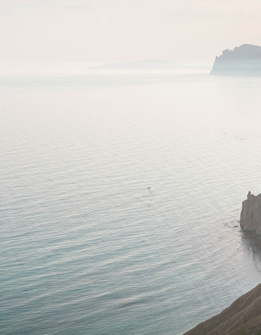 a hiker sits on mountaintop rocks looking out over vast water and mountains