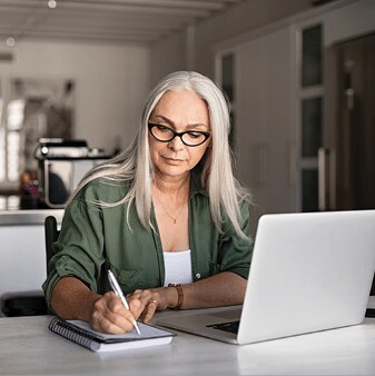 An older wman is working on a laptop. She is taking notes on a pad of paper.