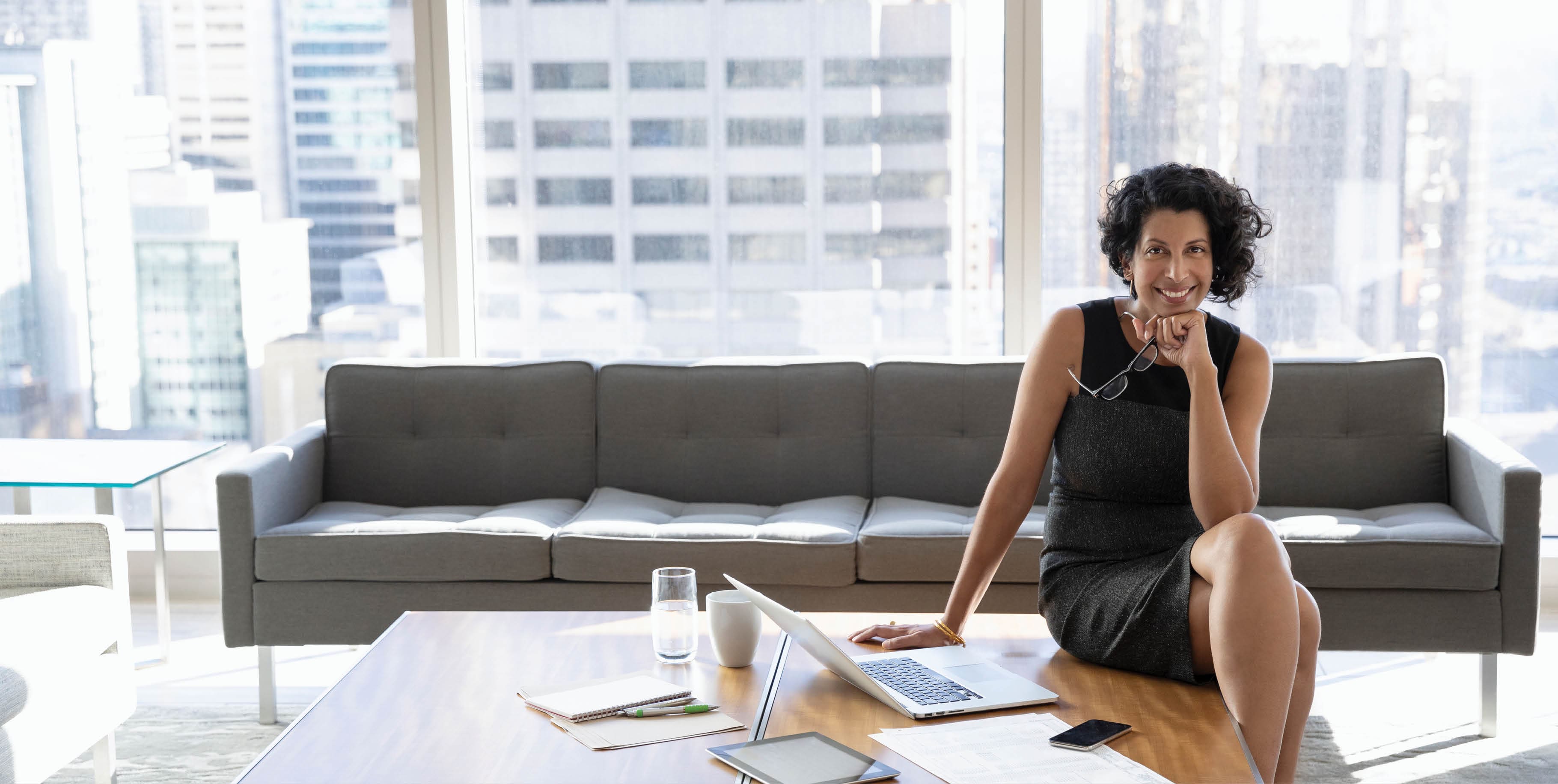 A smiling businesswoman is sitting on a desk next to a laptop, in a modern high rise office.