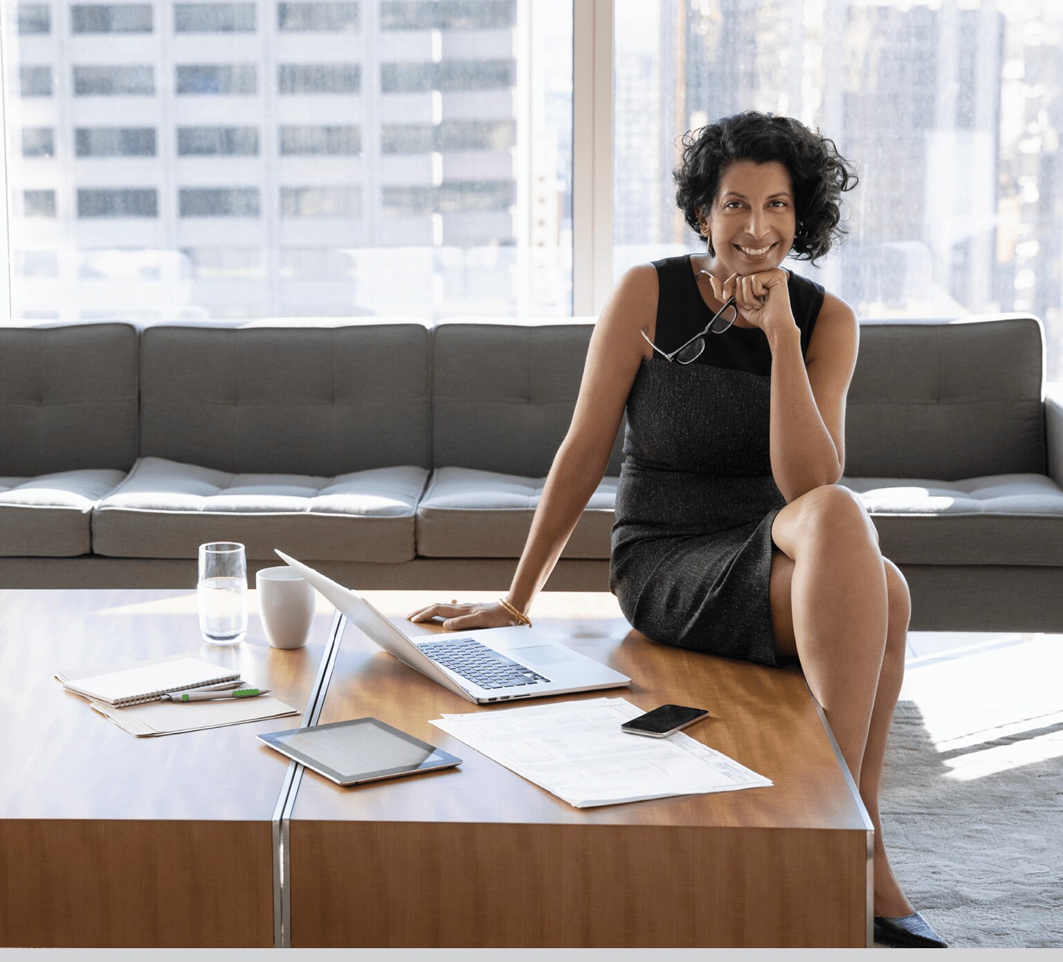 A smiling businesswoman is sitting on a desk next to a laptop, in a modern high rise office.