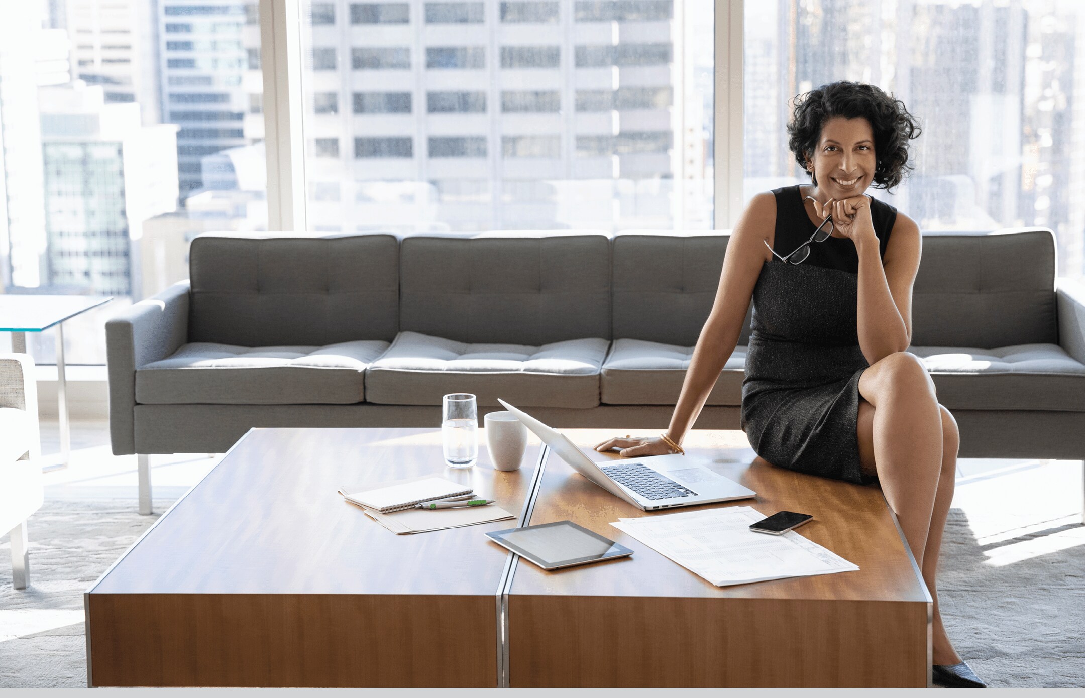 A smiling businesswoman is sitting on a desk next to a laptop, in a modern high rise office.