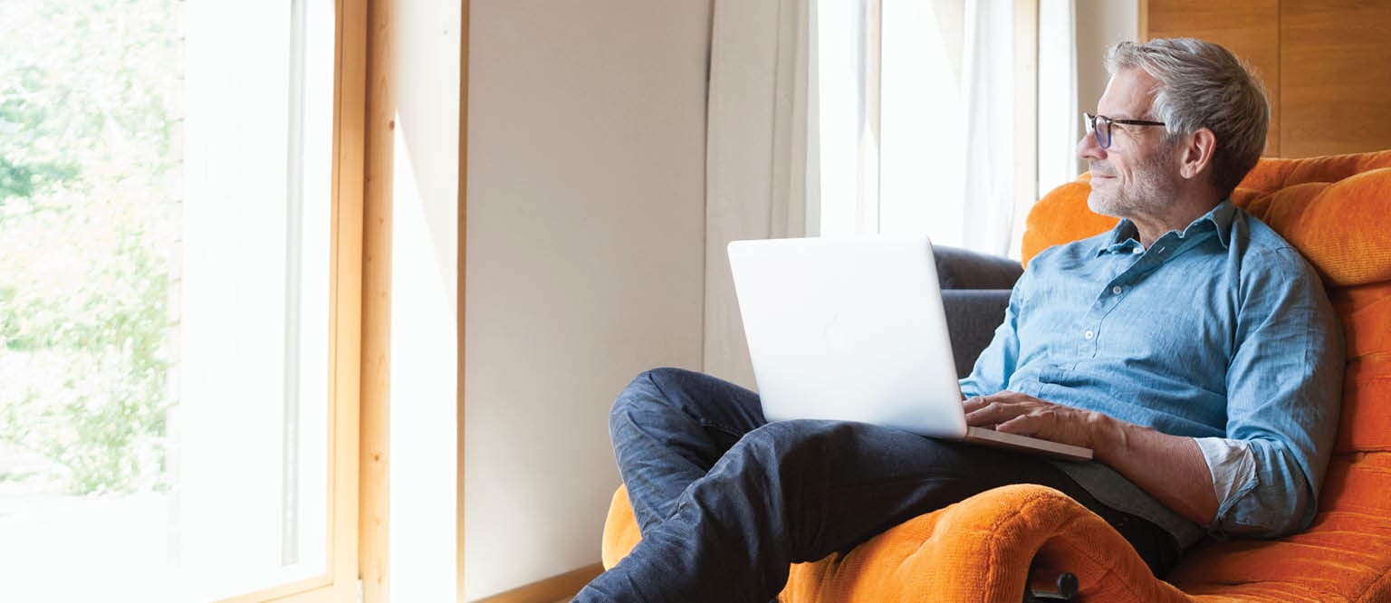 A middle-aged man is sitting with a laptop on his lap. He is looking out the window.