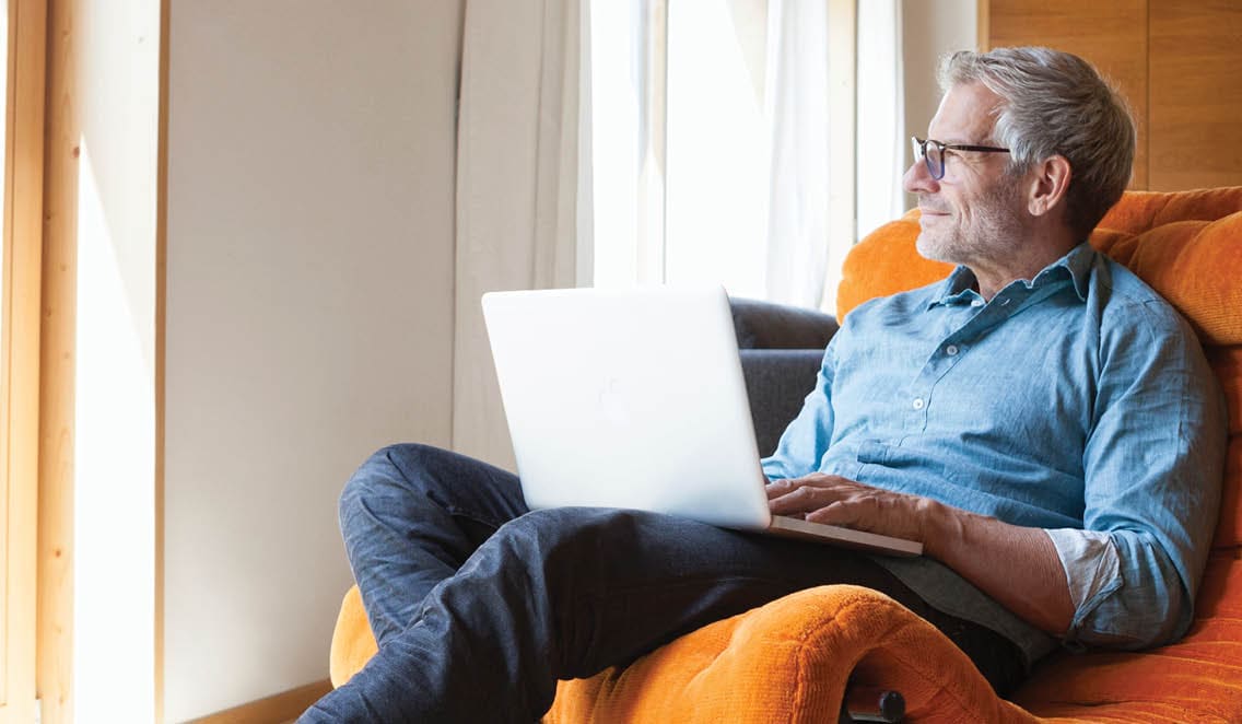 A middle-aged man is sitting with a laptop on his lap. He is looking out the window.