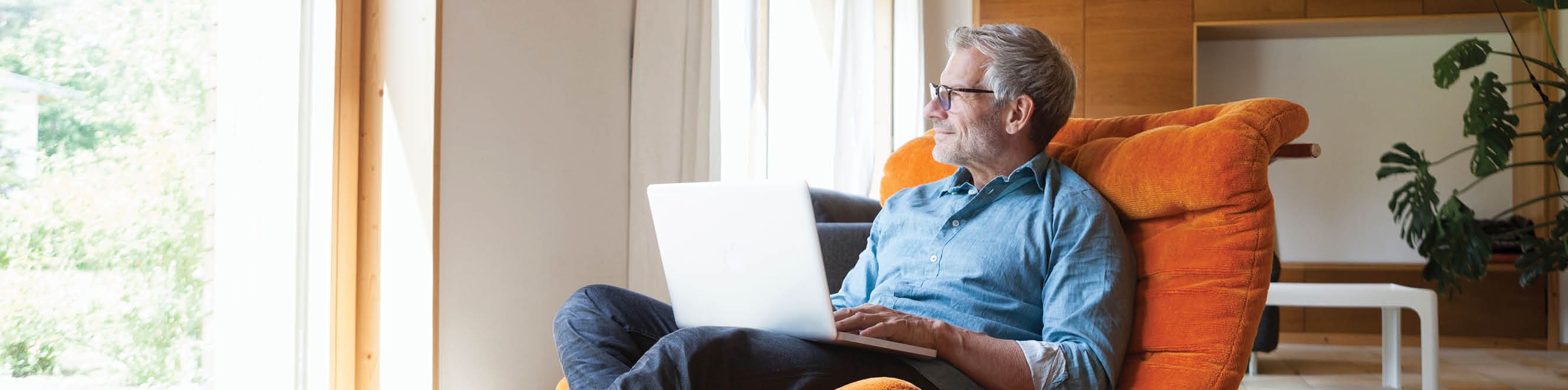 A middle-aged man is sitting with a laptop on his lap. He is looking out the window.