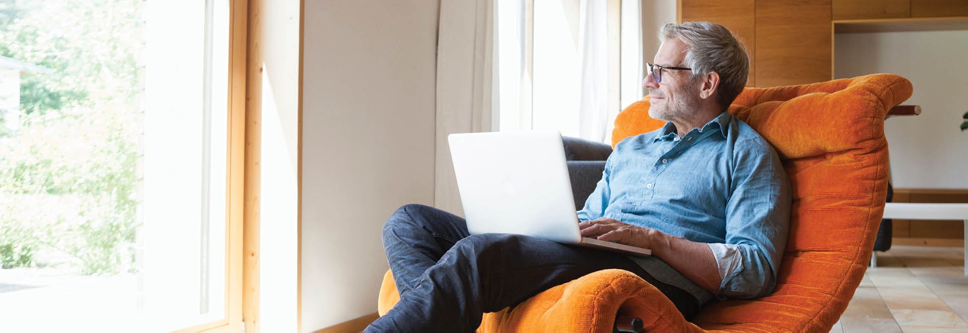 A middle-aged man is sitting with a laptop on his lap. He is looking out the window.