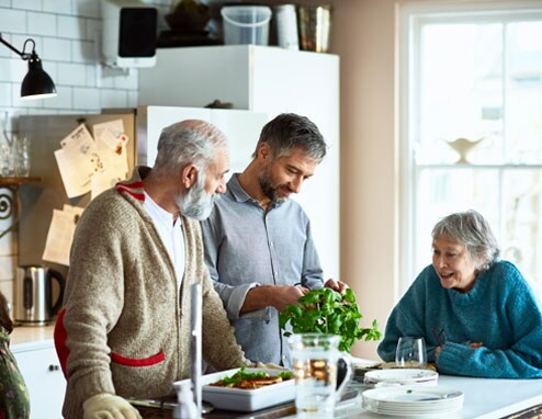 An elderly couple and their son are gather around a kitchen island. The son is helping his parents cook.