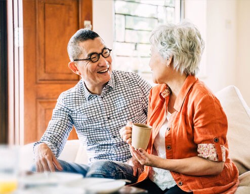 An older Asian couple is sitting on a couch. They are talking and laughing enjoying the relaxation of retirement.