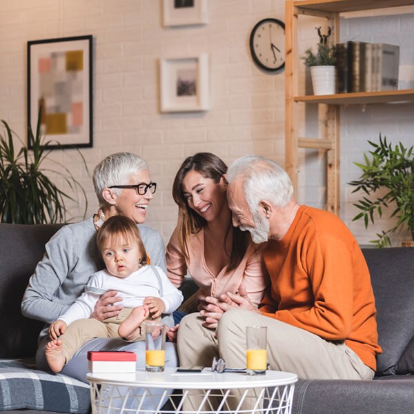 Grandparents are sitting on a couch with their daughter and grandaughter. They are engaged in conversation. The grandaughter in on the grandmother's knee. They are all smiling.