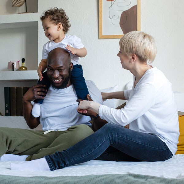 A mixed race couple is sitting on a bed. The father has their young daughter sitting on his shoulders. They are all smiling and laughing.