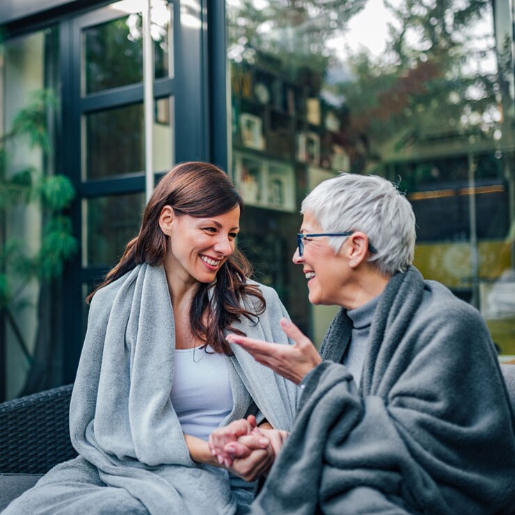 Women are more likely than men to become family caregivers. A younger female caregiver is sitting on a bench holding hands with an older woman. They are engaged in conversation and smiling.