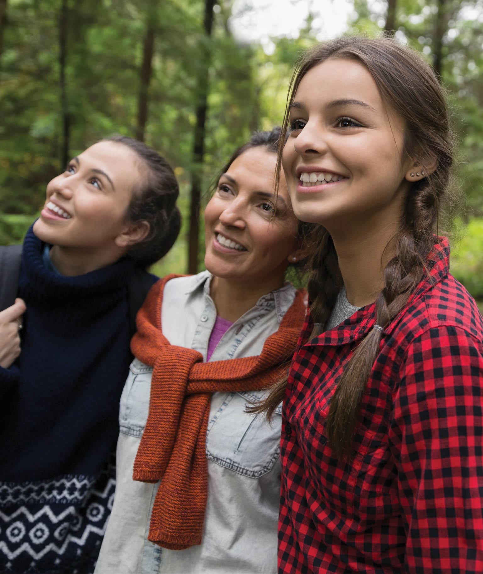 A mother and her two daughters are hiking through the wood. They are looking up in the trees and smiling.