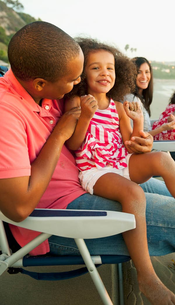 A mixed race couple are sitting in beach chairs with their two daughters. The father has one of the daughters on his lap. He is holding her shoulders and talking to her. Everyone is smiling.