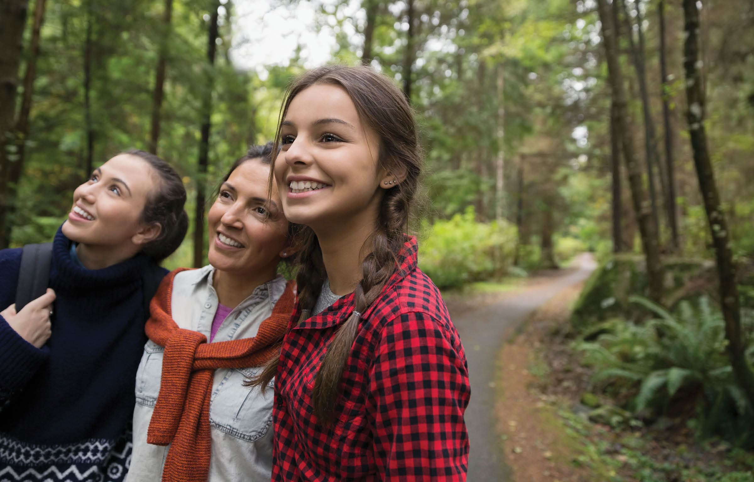 A mother and her two daughters are hiking through the wood. They are looking up in the trees and smiling.