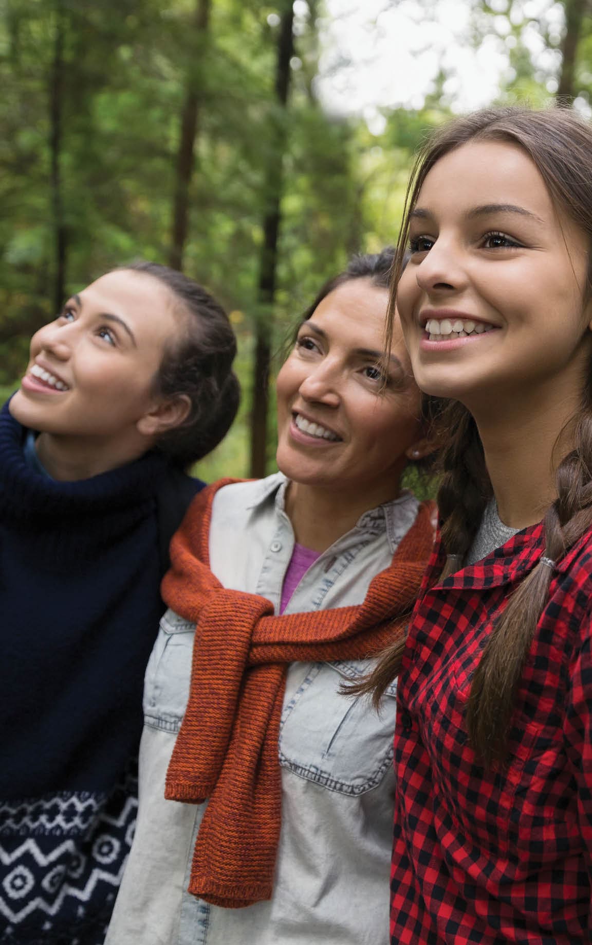 A mother and her two daughters are hiking through the wood. They are looking up in the trees and smiling.