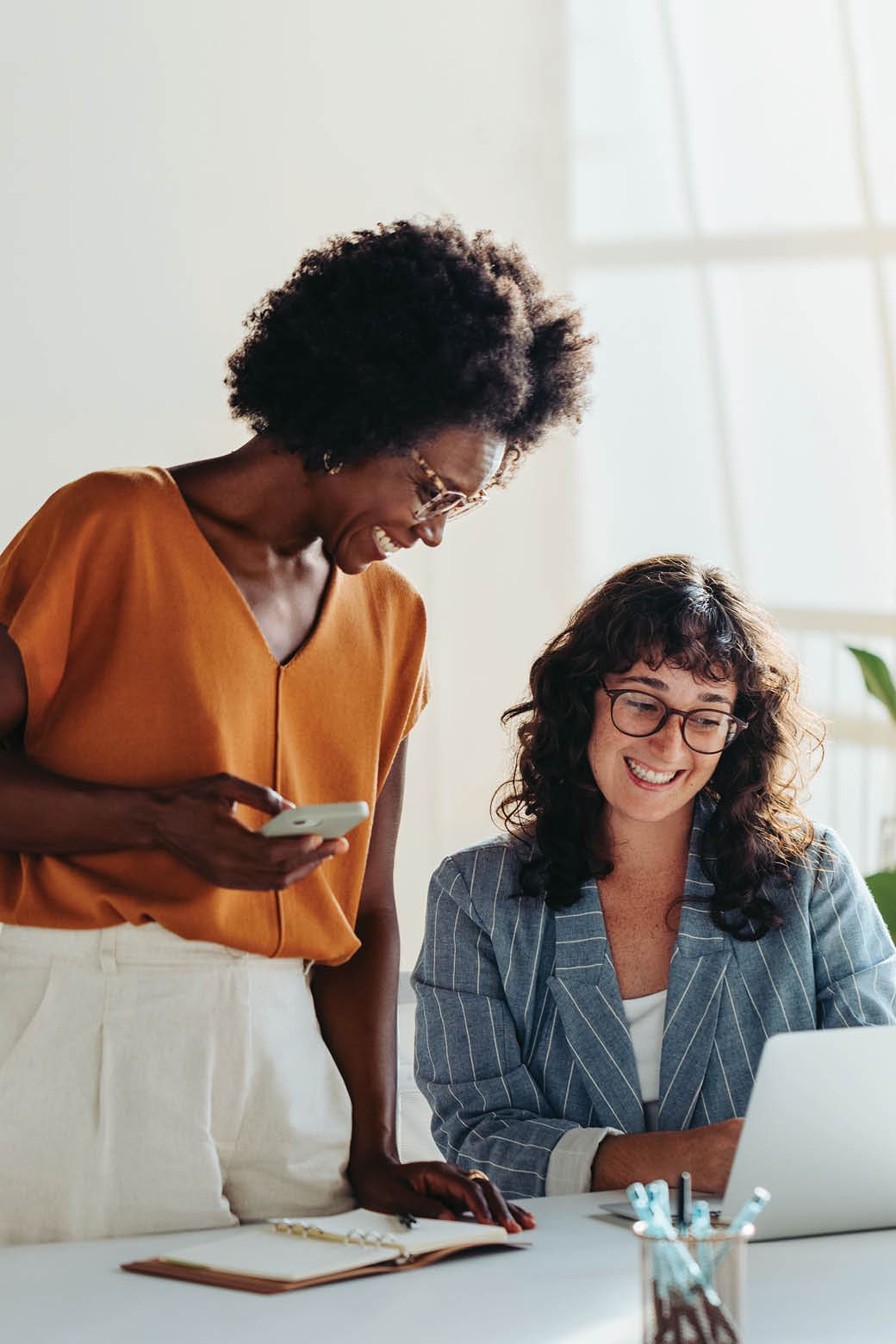 Happy professional women collaborating with enthusiasm in a modern office setting. Two female entrepreneurs using a laptop for a productive business meeting in a corporate workplace.