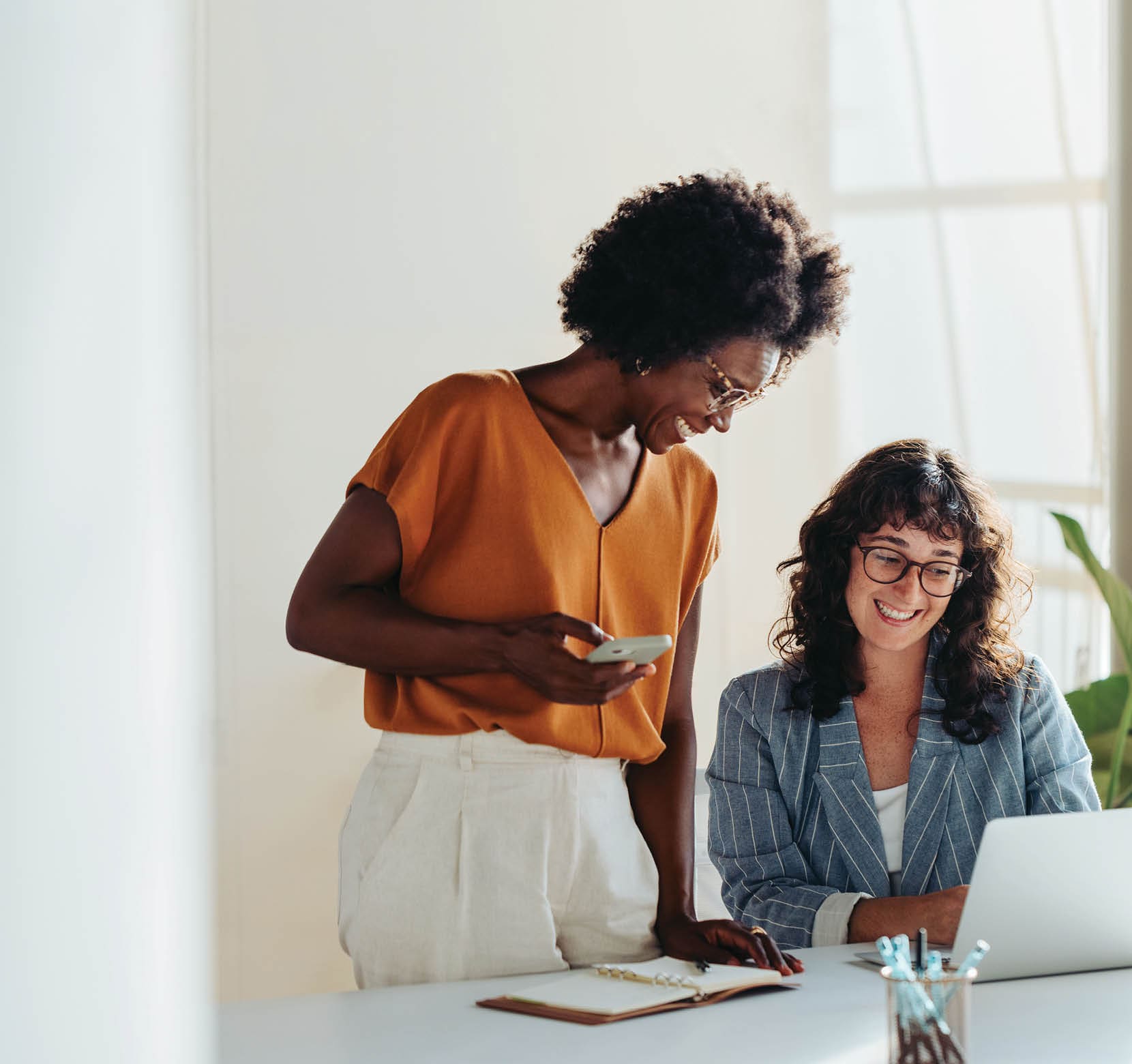 Happy professional women collaborating with enthusiasm in a modern office setting. Two female entrepreneurs using a laptop for a productive business meeting in a corporate workplace.
