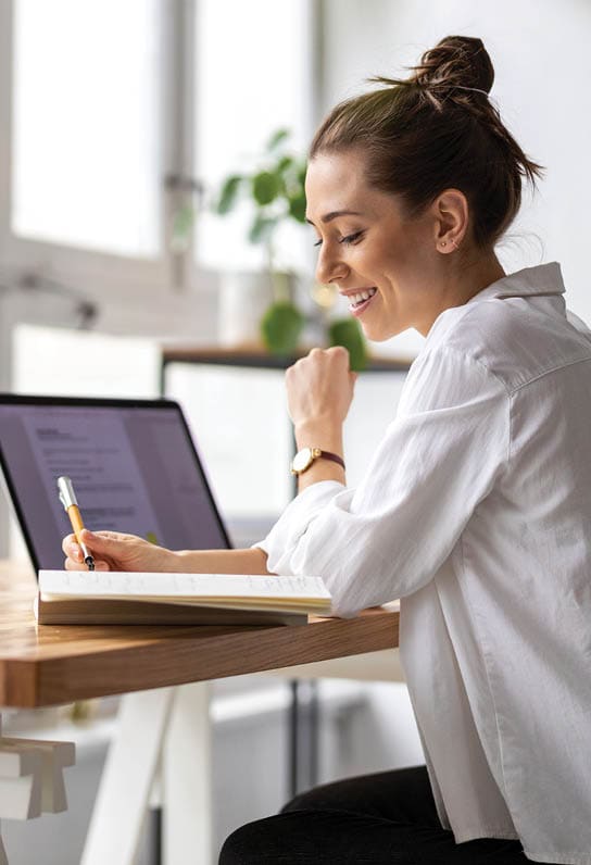 woman with laptop writing in notebook