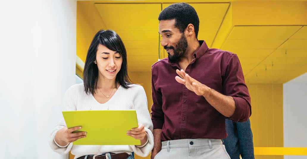 two colleagues looking at a printin out while walking down an office hall